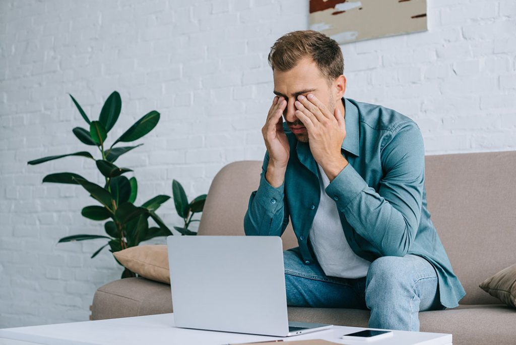 young man rubbing eyes while using with laptop at home after surgery